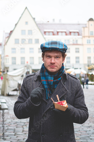 A handsome young man in a grey coat with a Scottish tweed cap and tartan scarf holding a dessert and standing on a Tallinn Town Hall Square on a winter day with a Christmas fare in the background