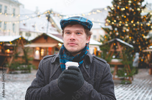 A handsome young man in a grey coat with a Scottish tweed cap and tartan scarf holding cocoa cup and standing on a Tallinn Town Hall Square with a Christmas tree and a Christmas fare in the background