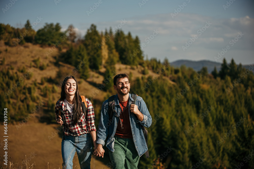 A diverse couple in their thirties, with beaming smiles, proudly hiking up a sun-drenched hillside with backpacks