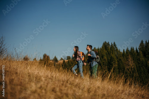 A diverse couple in their thirties, with beaming smiles, proudly hiking up a sun-drenched hillside with backpacks © La Famiglia