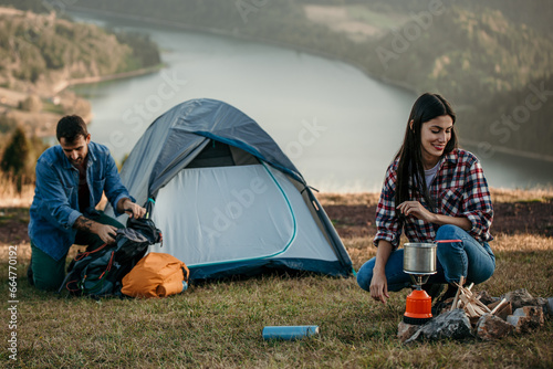 Multiethnic pair enjoying camping, making food on a camp stove, lake in the background.