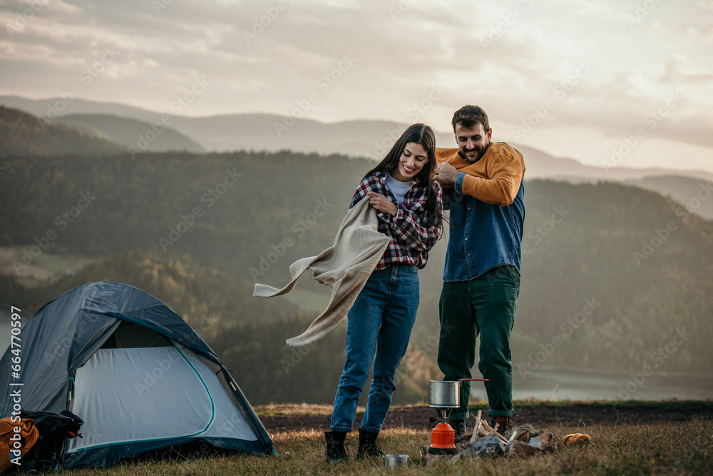 Fototapeta premium Diverse couple, putting on warm clothes, enjoy camping by a lake with a stove