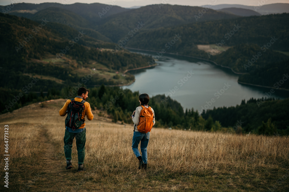 Two friends walking uphill with backpacks, catching sight of a serene lake