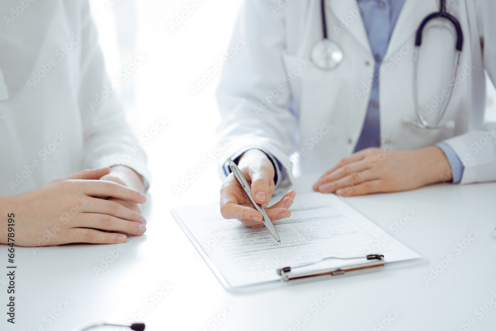 Doctor and patient sitting near of each other at the desk in clinic. The focus is on female physician's hands filling up the medication history record form or checklist, close up. Medicine concept Doctor and patient sitting near of each other at the desk in clinic. The focus is on female physician's hands filling up the medication history record form or checklist, close up. Medicine concept