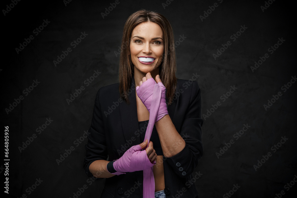Fototapeta premium Beautiful woman in a black jacket and sports bra, wrapping boxing wraps with mouthguard against a dark wall