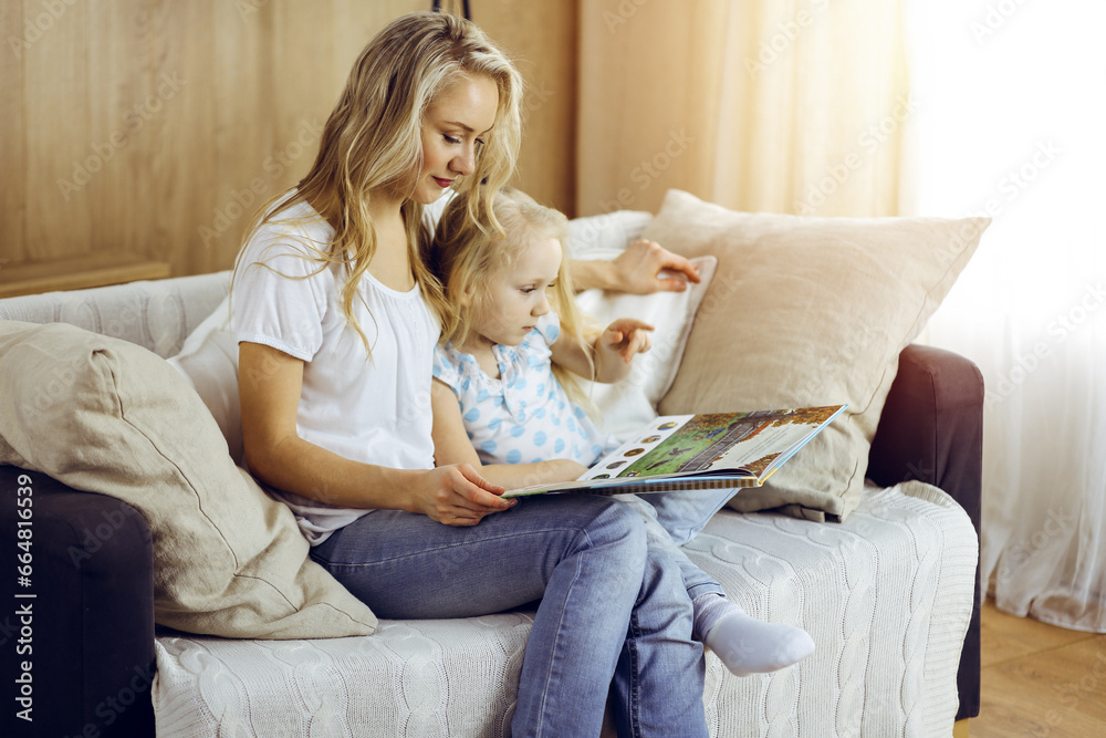 Happy family. Blonde young mother reading a book to her cute daughter while sitting at wooden floor in sunny room. Motherhood concept Happy family. Blonde young mother reading a book to her cute daughter while sitting at wooden floor in sunny room. Motherhood concept