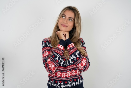 Curious Beautiful blonde girl wearing christmas sweater keeps hands under chin bites lips and looks with interest aside.