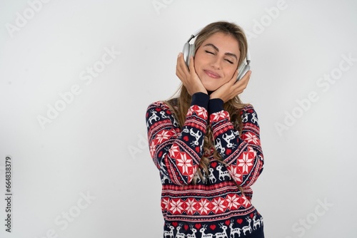 Pleased Beautiful blonde girl wearing christmas sweater enjoys listening pleasant melody keeps hands on stereo headphones closes eyes. Spending free time with music photo