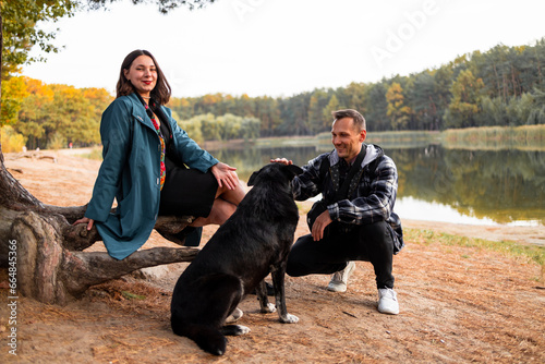 Attractive woman and handsome man spending time together with dog in autumn forest near lake. © Mykola