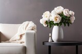 Vase of white peonies with coffee table and armchair near grey wall.