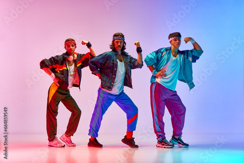 Strength. Three young men, friends wearing colorful old style sportswear, showing muscles against gradient pink blue background in neon light. Concept of sportive and active lifestyle, retro. Ad