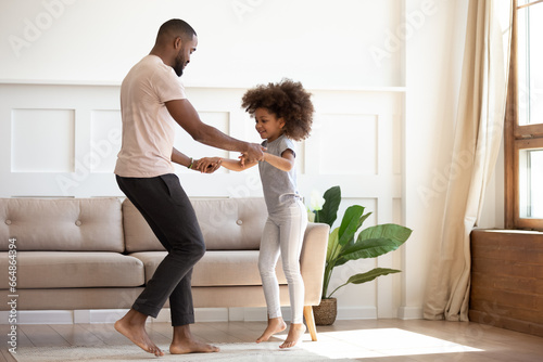 Happy African family father and cute little kid daughter jump having fun in living room in the morning, loving Black dad holding hands of child girl dancing together enjoy funny activity play at home