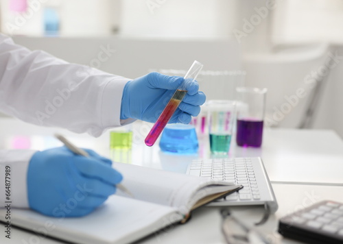 Employee of the chemical laboratory teacher chemist holds a silvery pen in his hand makes notes in the diary records. Test data from reactions examining test tubes with the substance arm in gloves.
