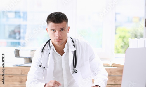 Handsome smiling male doctor sit at workplace in office. Physical and disease prevention patient aid exam visit 911 ward round prescribe remedy healthy lifestyle consultant profession concept