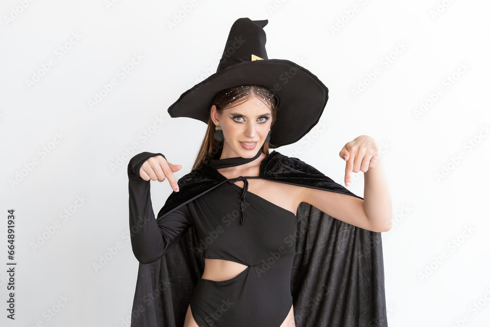 Attractive young woman in black clothes and black hat points her fingers down. White background. Halloween party. Attractive young woman in black clothes and black hat points her fingers down. White background. Halloween party.