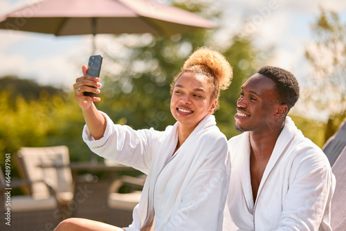 Couple In Robes Outdoors On Loungers By Pool On Spa Day Posing For Selfie On Mobile Phone © Monkey Business