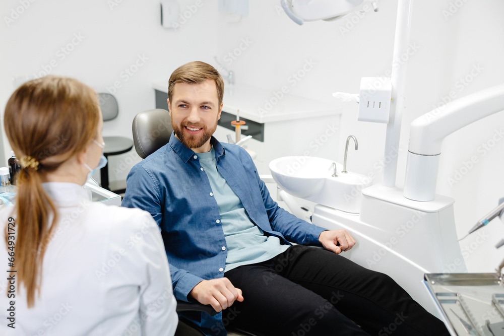 Blonde female dentist in dental office talking with male patient and preparing for treatment. Handsome bearded man in dentist chair looking at his doctor with smile Blonde female dentist in dental office talking with male patient and preparing for treatment. Handsome bearded man in dentist chair looking at his doctor with smile