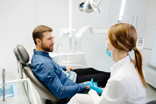 Blonde female dentist in dental office talking with male patient and preparing for treatment. Handsome bearded man in dentist chair looking at his doctor with smile photo
