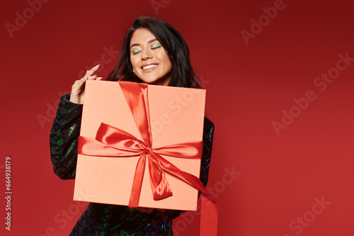 pleased asian woman in green jacket with sequins holding wrapped Christmas present on red backdrop