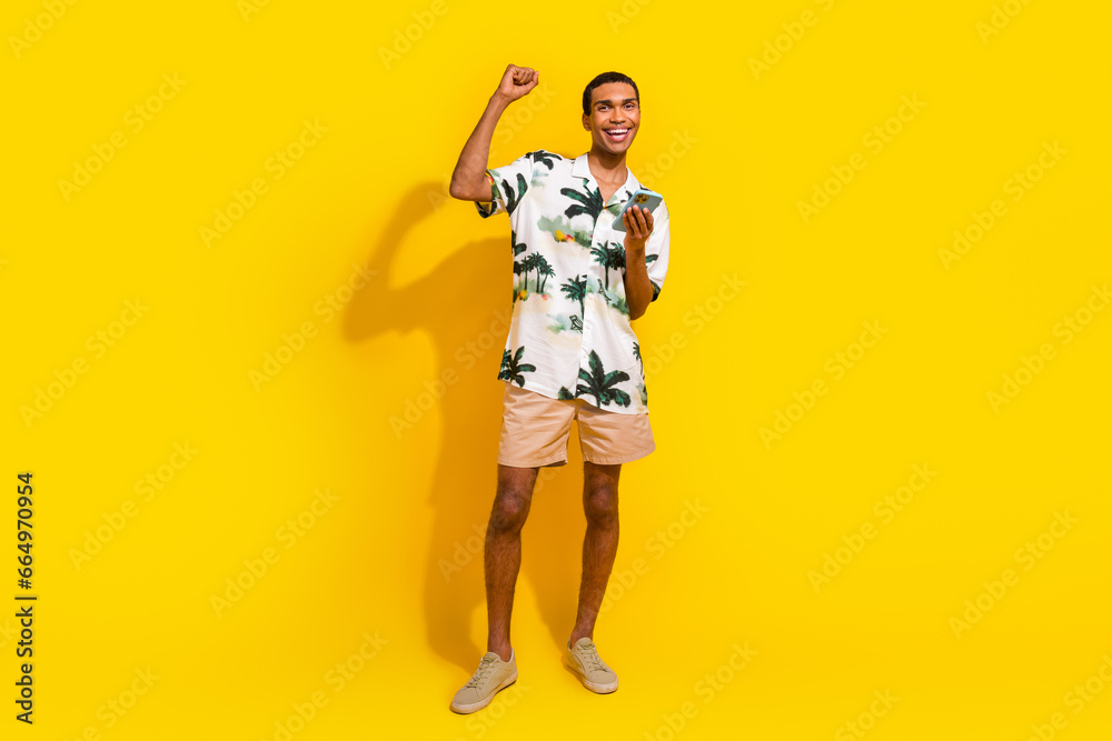 Full body photo of attractive young man hold device raise fist wear trendy palms print clothes isolated on yellow color background Full body photo of attractive young man hold device raise fist wear trendy palms print clothes isolated on yellow color background