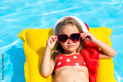 Cute happy little girl in Santa Claus hat, sunglasses and red swimsuit lying on inflatable mattress in swimming pool on tropical vacation. Christmas in warm countries. Advertising for tour operators