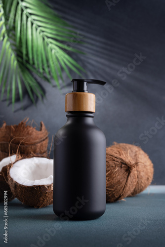Black dispenser bottle and fresh coconut on dark background with palm leaves