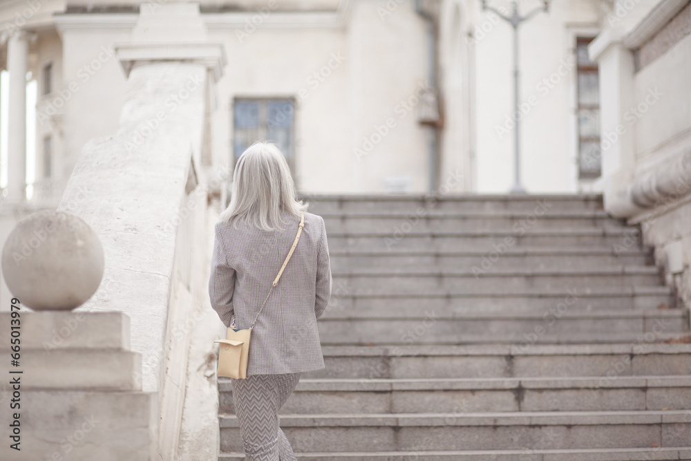 Back view of a  beauttiful woman with grey hair is walking arounfd the old town. A stylidh woman is wearing a gray coat, a skirt and a bag,