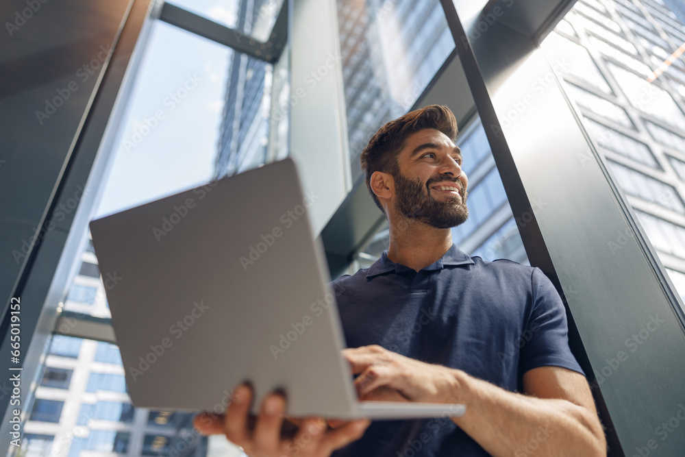 Handsome freelancer with laptop standing in coworking near window and looks away Handsome freelancer with laptop standing in coworking near window and looks away