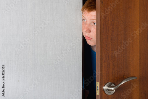 A teenage boy with red hair peeks curiously from behind the door of the room.