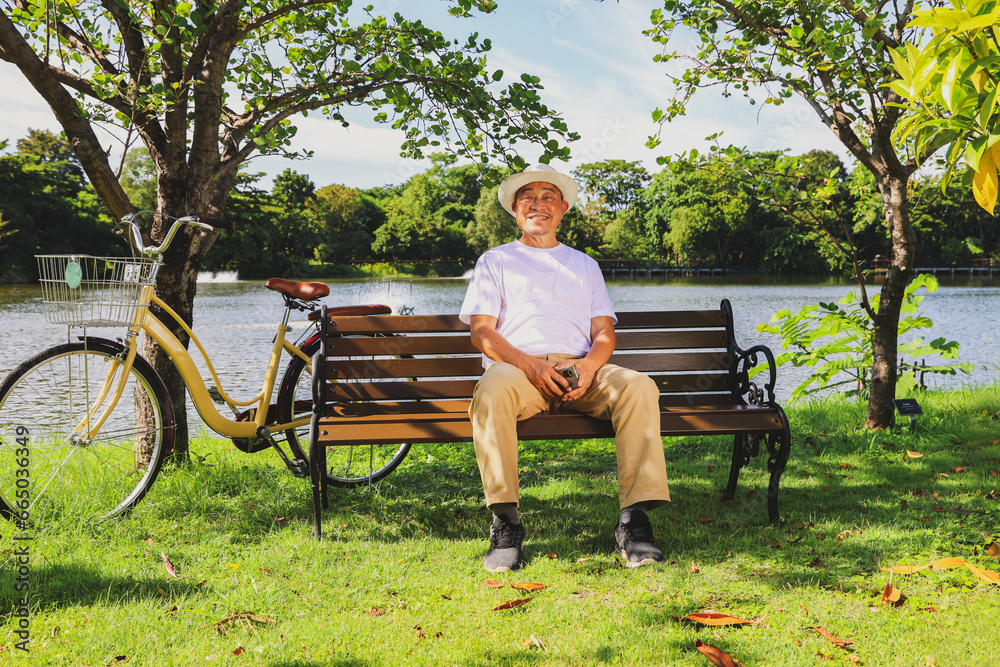 Senior man wearing sunshade asian hat healthy and good mood sitting on a bench under tree in the park with bicycle riding doing exercise for health clear shady day looking at the camera smiling happy.