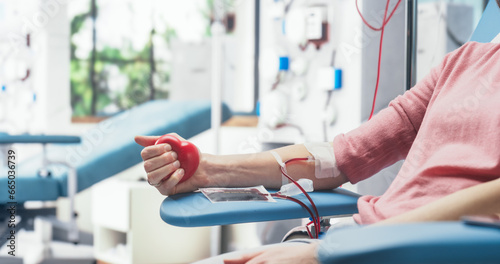 Close Up Shot Of Hand Of Female Blood Donor With an Attached Catheter. Caucasian Woman Squeezing Heart-Shaped Red Ball To Pump Blood Through The Tubing Into Bag. Donation For Victims Of Accidents.