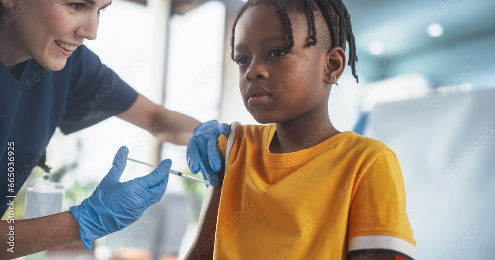 Young African American Boy Sitting In The Chair In Bright Hospital And Getting Polio Vaccine. Caucasian Female Nurse Performing Injection. Professional Woman Talks To Worried Kid, Calms Him Down. - obrazy, fototapety, plakaty 