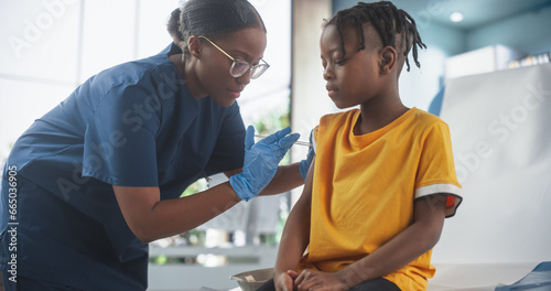 Young African American Boy Sitting In The Chair In Bright Hospital And Getting His Polio Vaccine. Female Black Nurse Is Performing Injection. Professional Woman Talks To Worried Kid And Calms Him Down