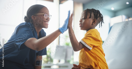 Young African American Boy Sitting In The Chair In Bright Hospital And Getting His Flu Vaccine. Female Black Nurse Is Finished Performing Injection. Professional Woman High-Fives A Kid For Being Brave