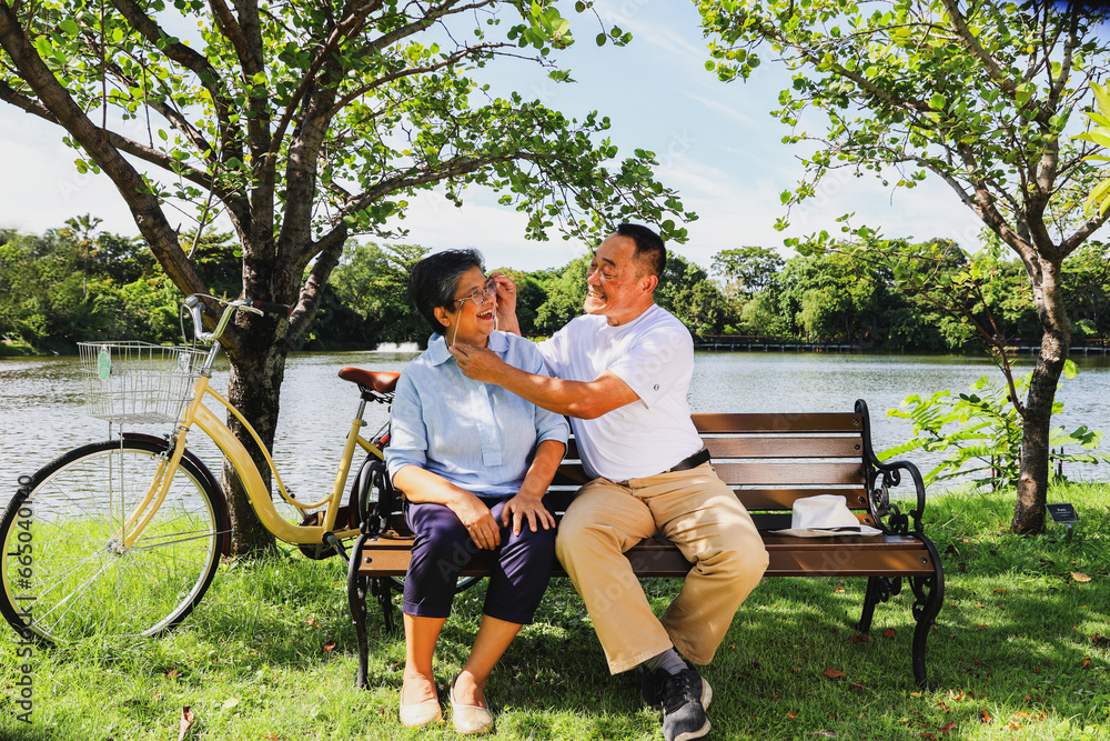 Fototapeta premium Senior asian retirement couple sits and admires the sunny shady outdoor garden : Husband takes care each other shows concern for his wife's eye health and helps to steady her eyeglasses on her face.