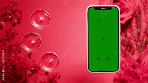 Phone with green screen chroma key on red and pink background close up, top view. Smartphone on summer bright texture studio shot. Composition of rose flowers, aroma and lipstick make photo