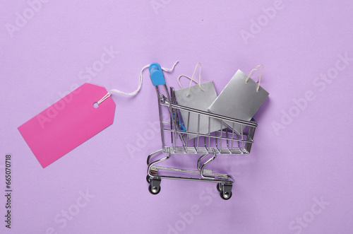 Miniature shopping bags with sale tag in supermarket trolley on purple pastel background. Black Friday, world shopping day, sale