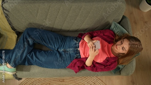 Top view capturing a young woman laying on the couch, looking at the screen of his smartphone. She is texting, chatting with someone online and smiling. photo