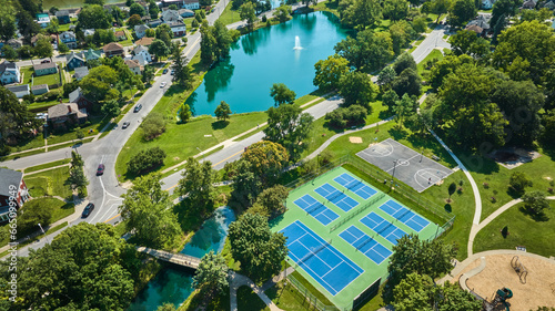 Fototapeta Naklejka Na Ścianę i Meble -  Lakeside Park bridge near tennis courts and basketball court with neighborhood and pond aerial