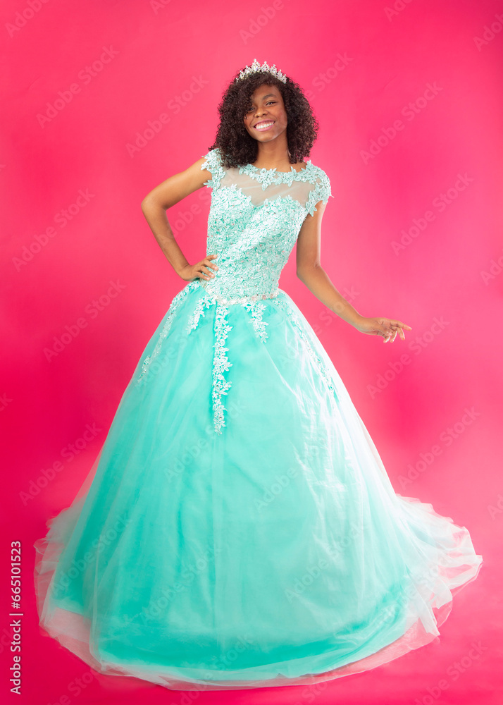 Cute hispanic girl celebrating her 16th birthday with a blue big dress in a pink background 