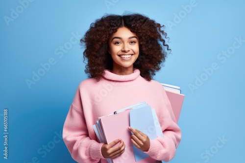 A young woman with a cheerful smile holding a stack of books in a cozy pink sweater