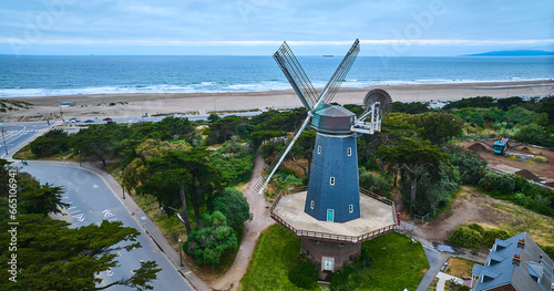 Blue murphy windmill surrounded by trees and roads overlooking blue ocean with sandy shore aerial