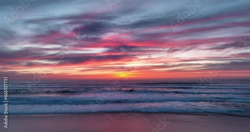 Aerial view of colorful sunset reflecting over the ocean, casting a warm and vibrant glow on the serene sandy beach below, during the stunning sundown.