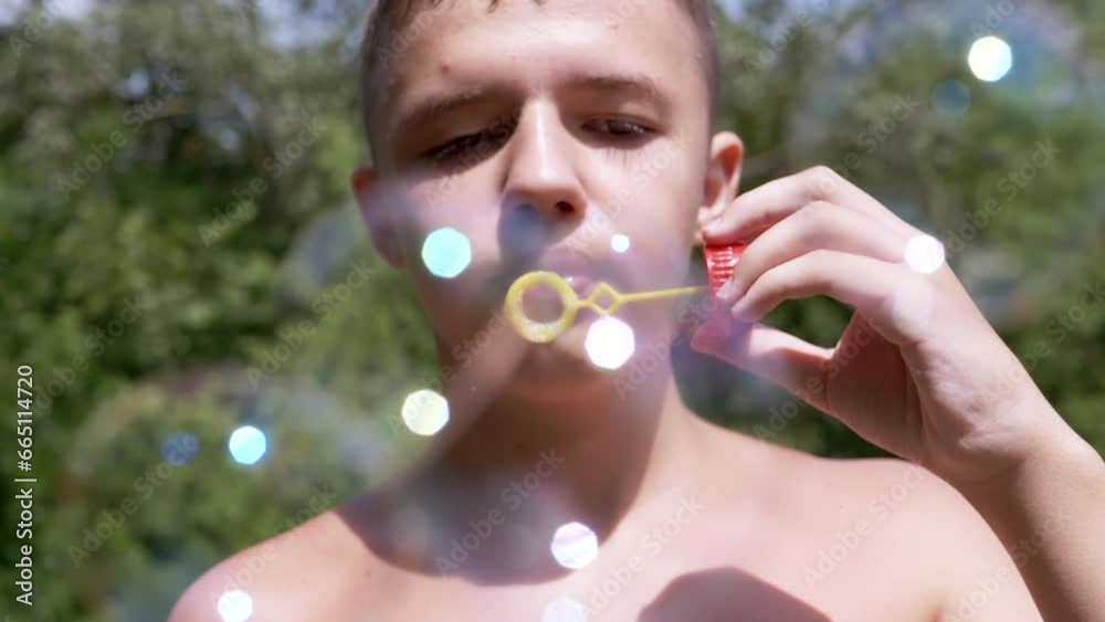 Boy with a Naked Torso Blowing Soap Bubbles in Nature in the Rays of Sunlight. Close up. A teenager is relaxing outdoors in the forest, blowing colorful bright soap bubbles. Blurred green background. Boy with a Naked Torso Blowing Soap Bubbles in Nature in the Rays of Sunlight. Close up. A teenager is relaxing outdoors in the forest, blowing colorful bright soap bubbles. Blurred green background.