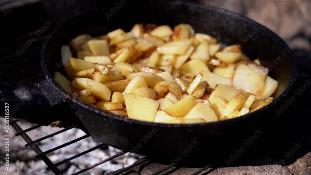 Cooking Fried Potatoes in Iron Frying Pan on a Campfire in the Forest Outdoors. Crispy pieces of potatoes with smoke on coals in the sunlight at sunset. Concept of food, picnic recreation in nature. Cooking Fried Potatoes in Iron Frying Pan on a Campfire in the Forest Outdoors. Crispy pieces of potatoes with smoke on coals in the sunlight at sunset. Concept of food, picnic recreation in nature.