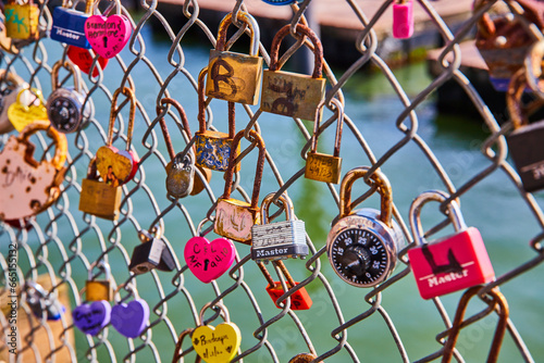 Photography Variety of love and heart shaped locks on chain link fence with water in backgro