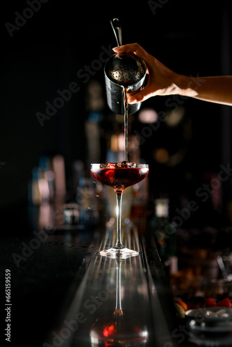 Close up view of woman bartender pours fine jet of red cocktail into drinking glass with ice standing on the bar counter