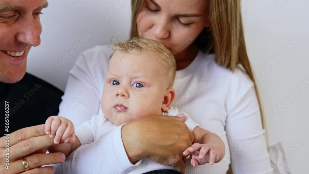 Caucasian woman holding her beautiful blond blue-eyed baby. Mom kisses her infant on the head and dad holds kid's hand. Caucasian woman holding her beautiful blond blue-eyed baby. Mom kisses her infant on the head and dad holds kid's hand.