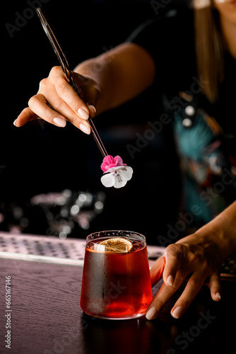 close-up of female bartender's hand holding tweezers with small fresh flower and decorating glass with brown alcoholic cocktail