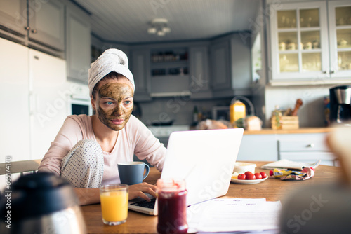 Attractive young woman with a facial mask using a laptop in the kitchen